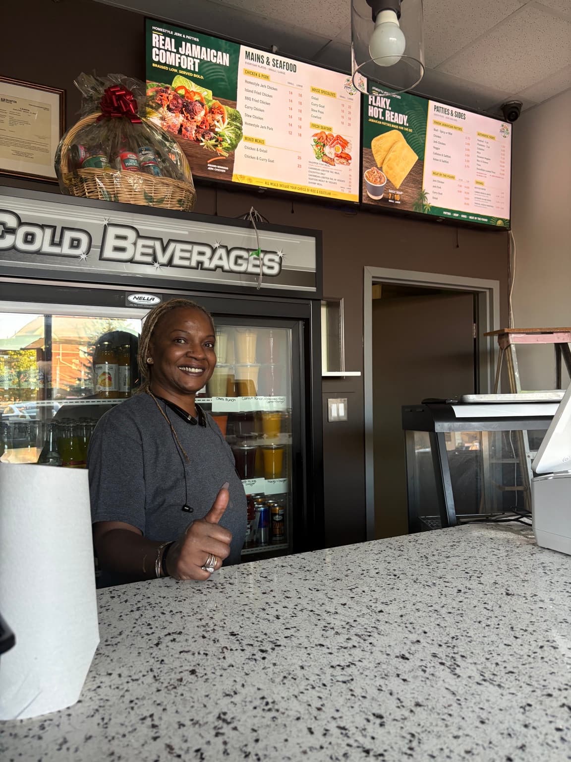 A woman giving a thumbs-up beneath HomeStyle Jerk & Patties digital menu boards at the counter